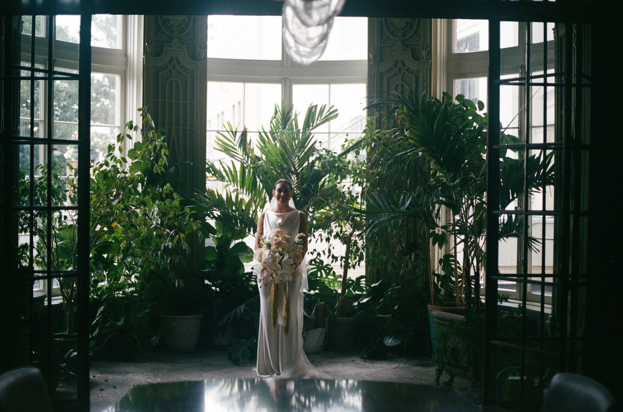 A bride standing among tall palms in a glass conservatory, holding a cascading bouquet.