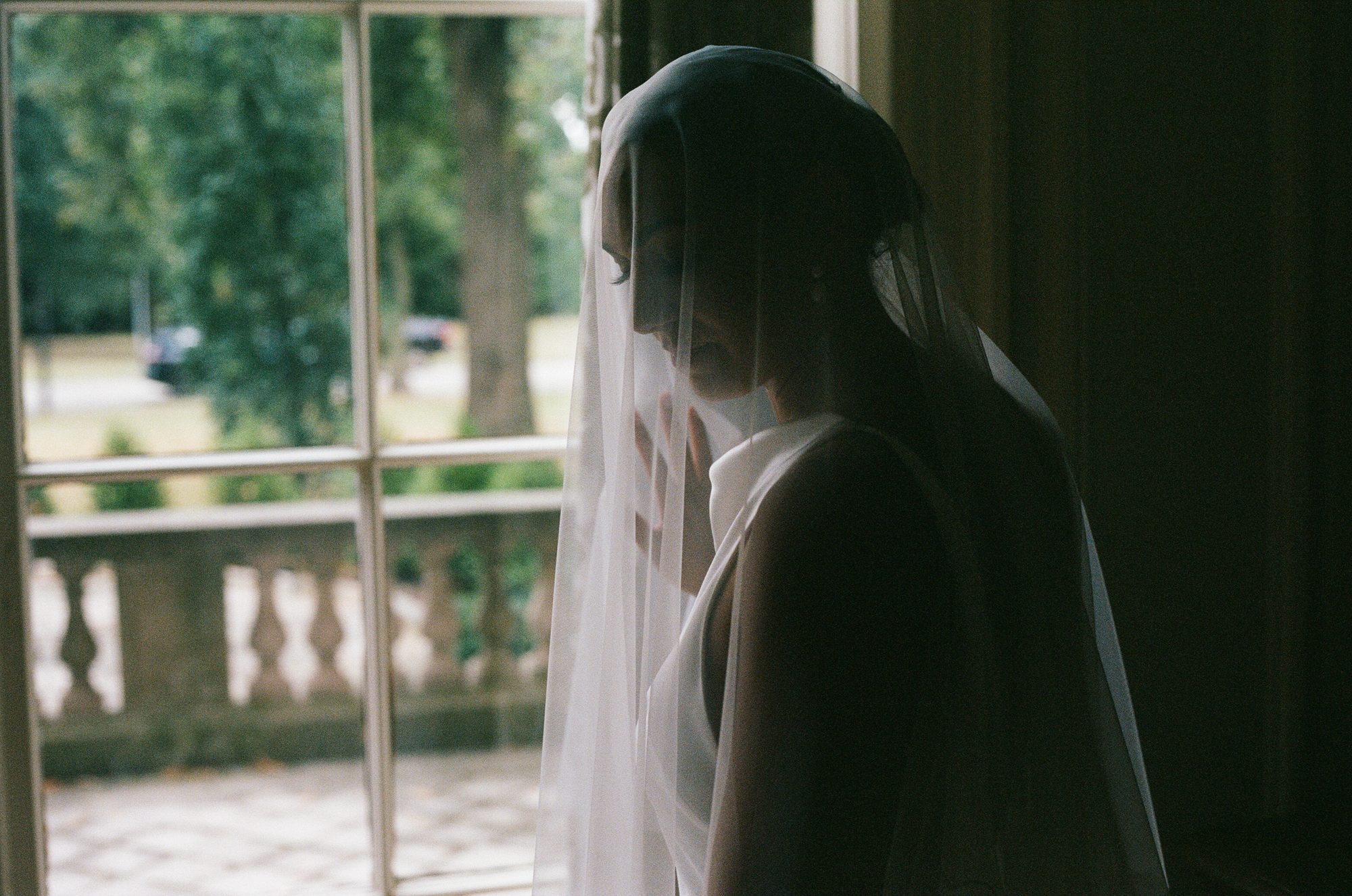 A bride in silhouette wearing a veil, looking down toward a window with a balustrade visible outside.