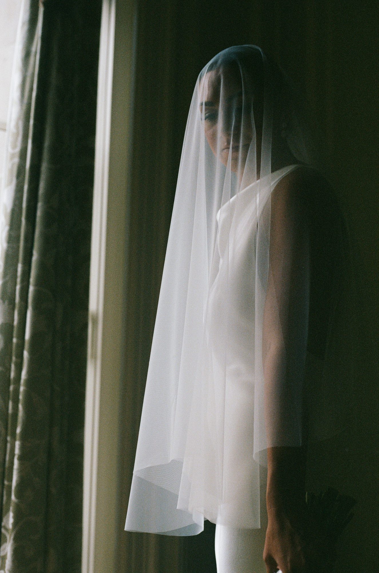 A bride in profile beneath a veil, standing against heavy curtains, dim natural light.
