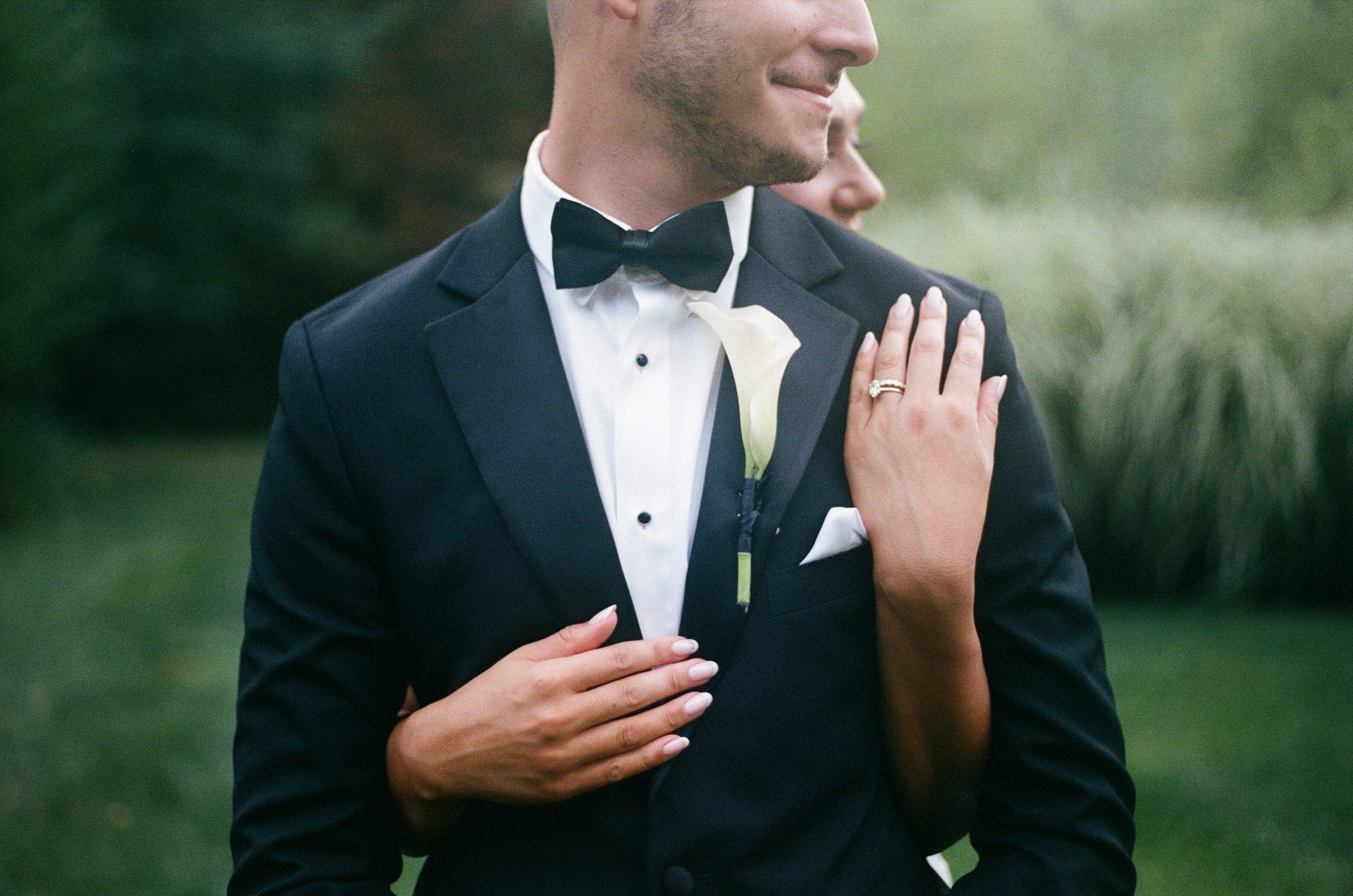A groom smiling in profile with a calla lily boutonniere, his partner's hand resting on his lapel, soft green background.