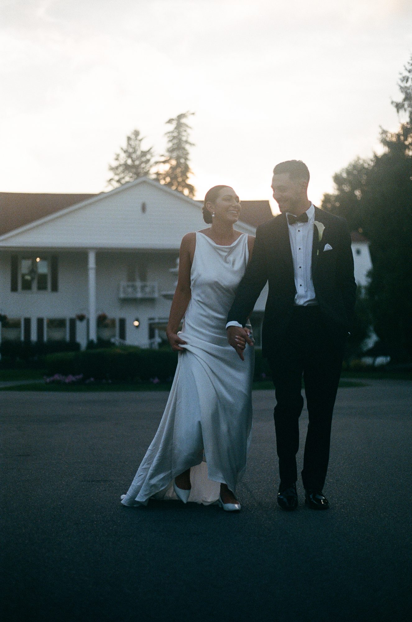 A bride and groom walking hand in hand across a driveway at dusk in front of a large white house.