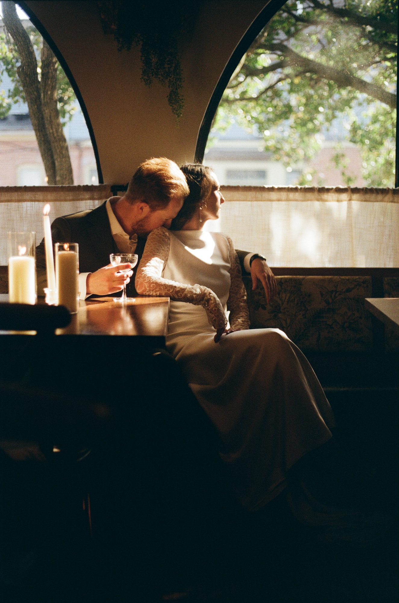 A couple leaning into each other in a candlelit restaurant booth, a glass of wine on the table, trees visible through the window.