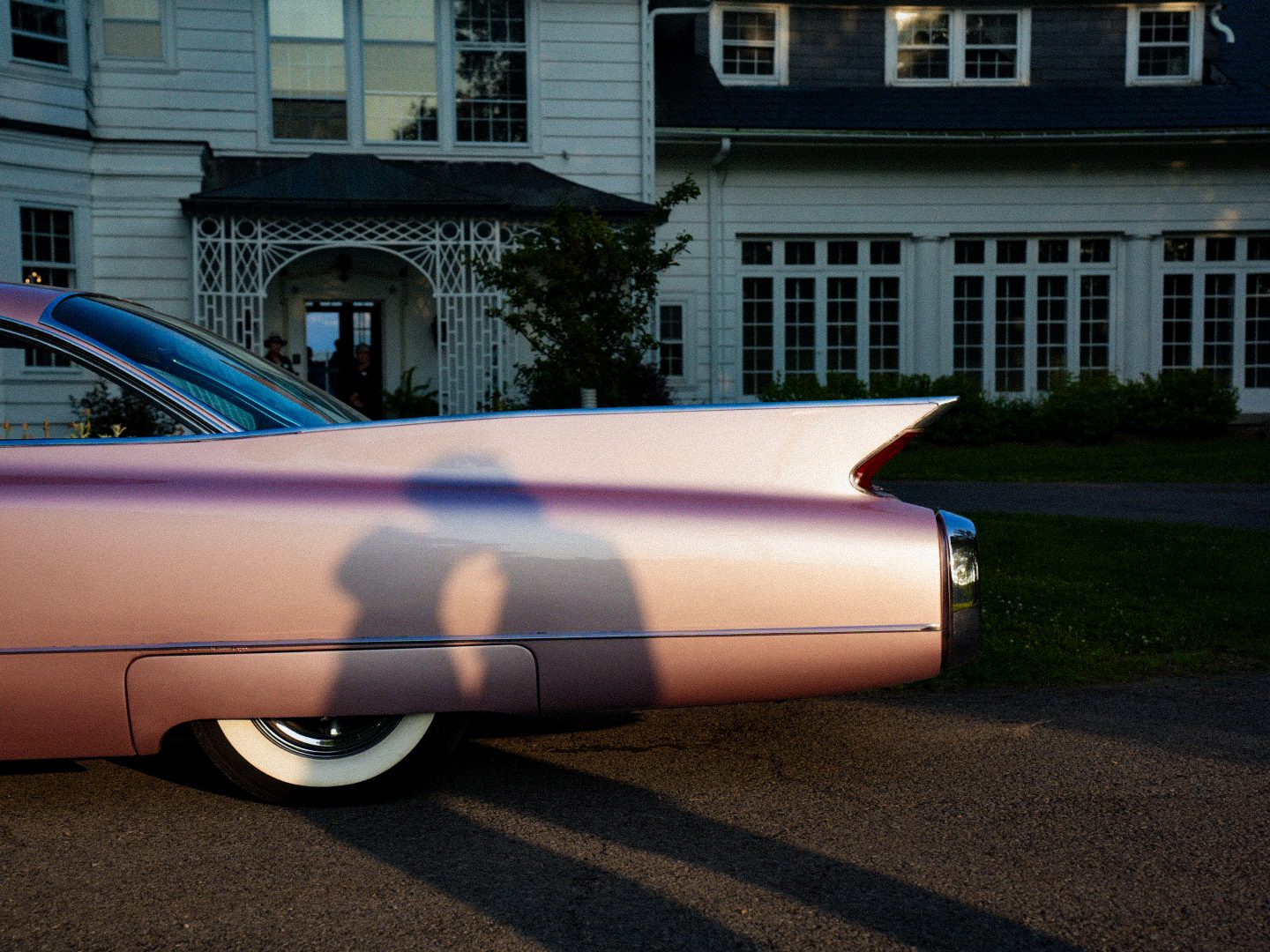 A vintage pink Cadillac with tailfins parked outside a white house, a couple's kissing silhouette cast across its side panel.