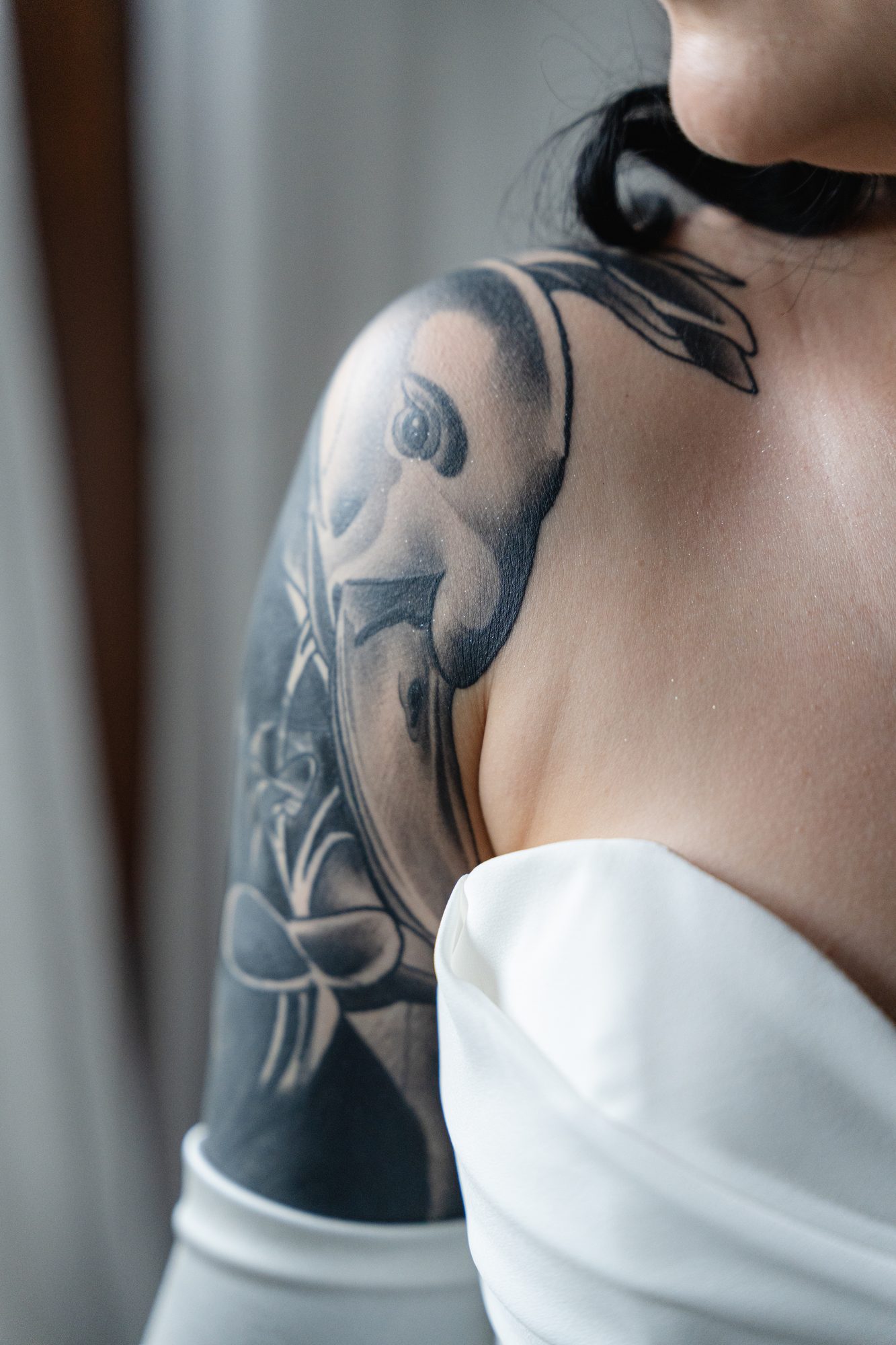 Close detail of a tattoo sleeve on a bride's shoulder as she adjusts her dress near a window.