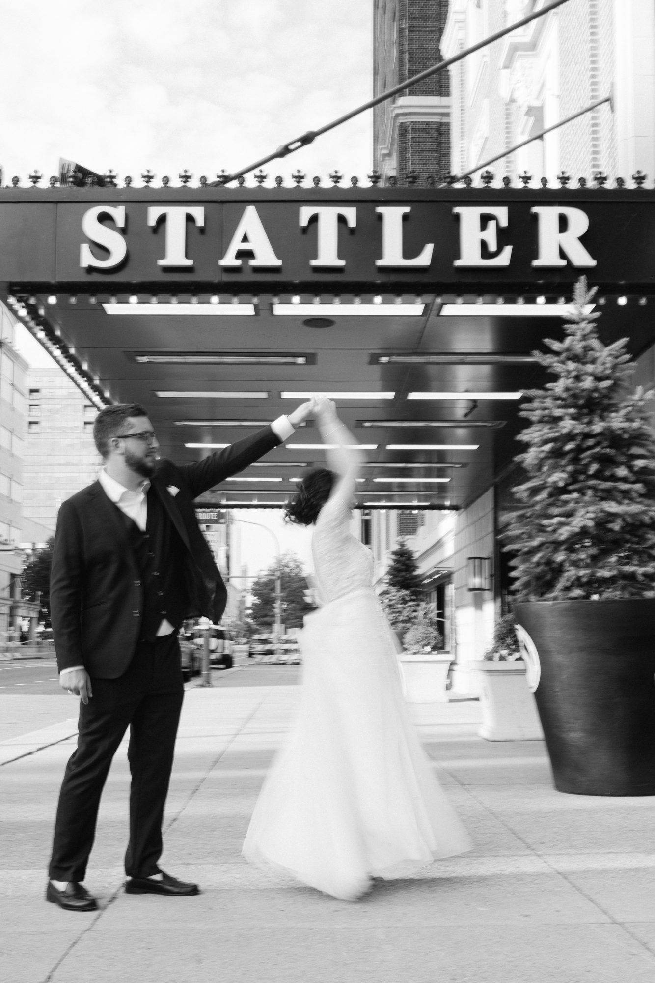 A groom twirling his bride under a hotel marquee, black and white.