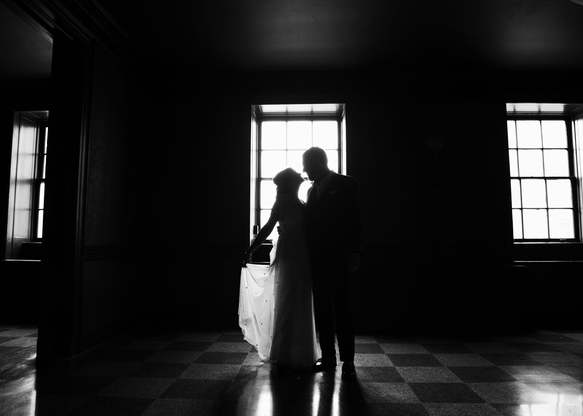 A bride and groom silhouetted in a dark room in front of a bright window, checkered floor below, black and white.