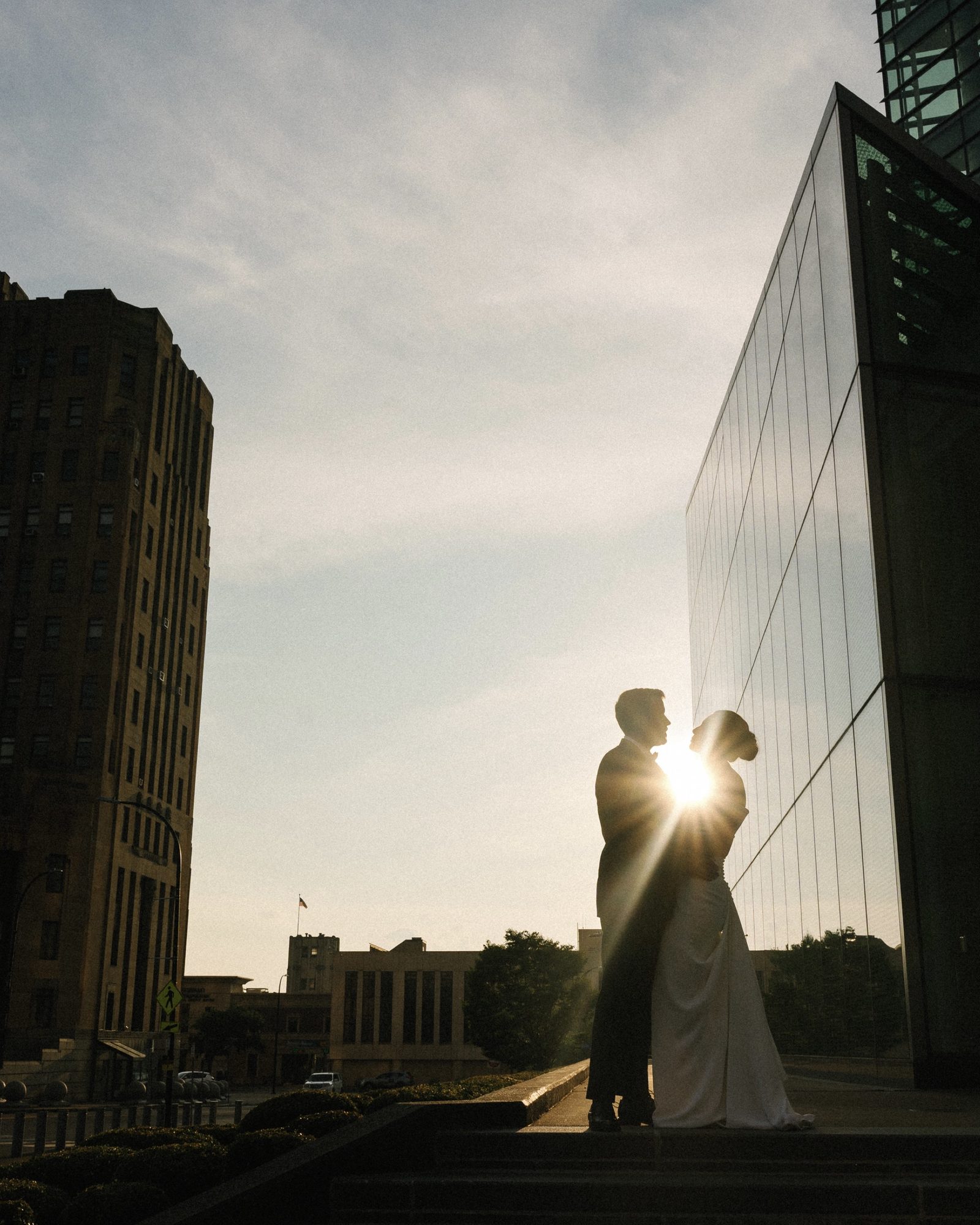 A bride and groom kissing, silhouetted against a glass building with the sun breaking between them.