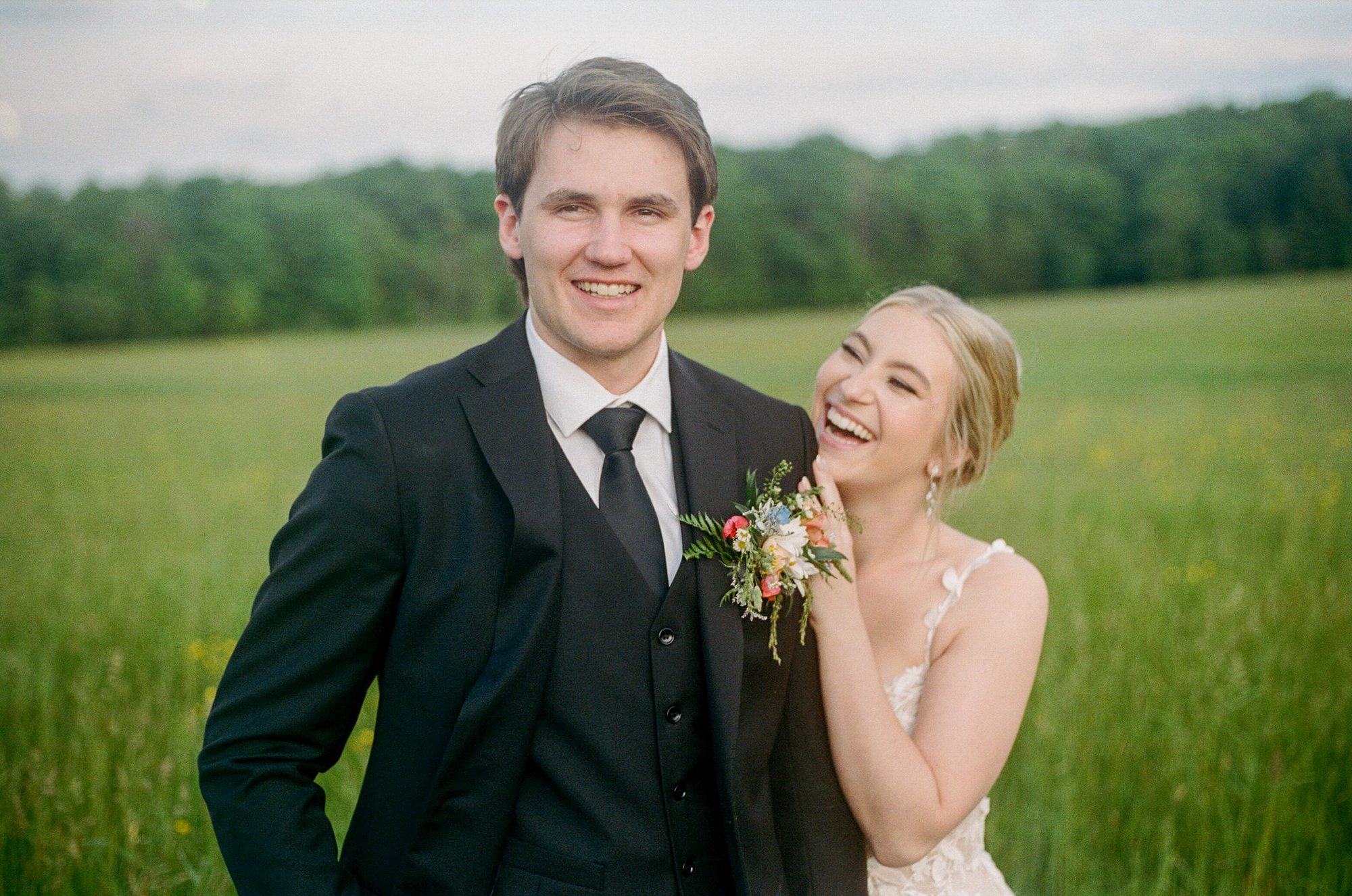 A groom laughing while his bride rests a hand on his shoulder, in front of a green field.
