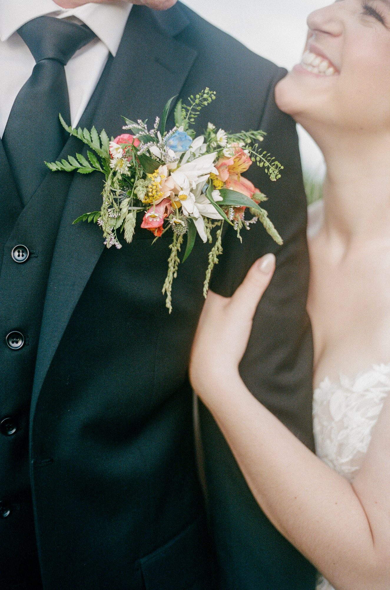 Close detail of a wildflower boutonniere on a groom's black suit jacket, bride laughing in the background.