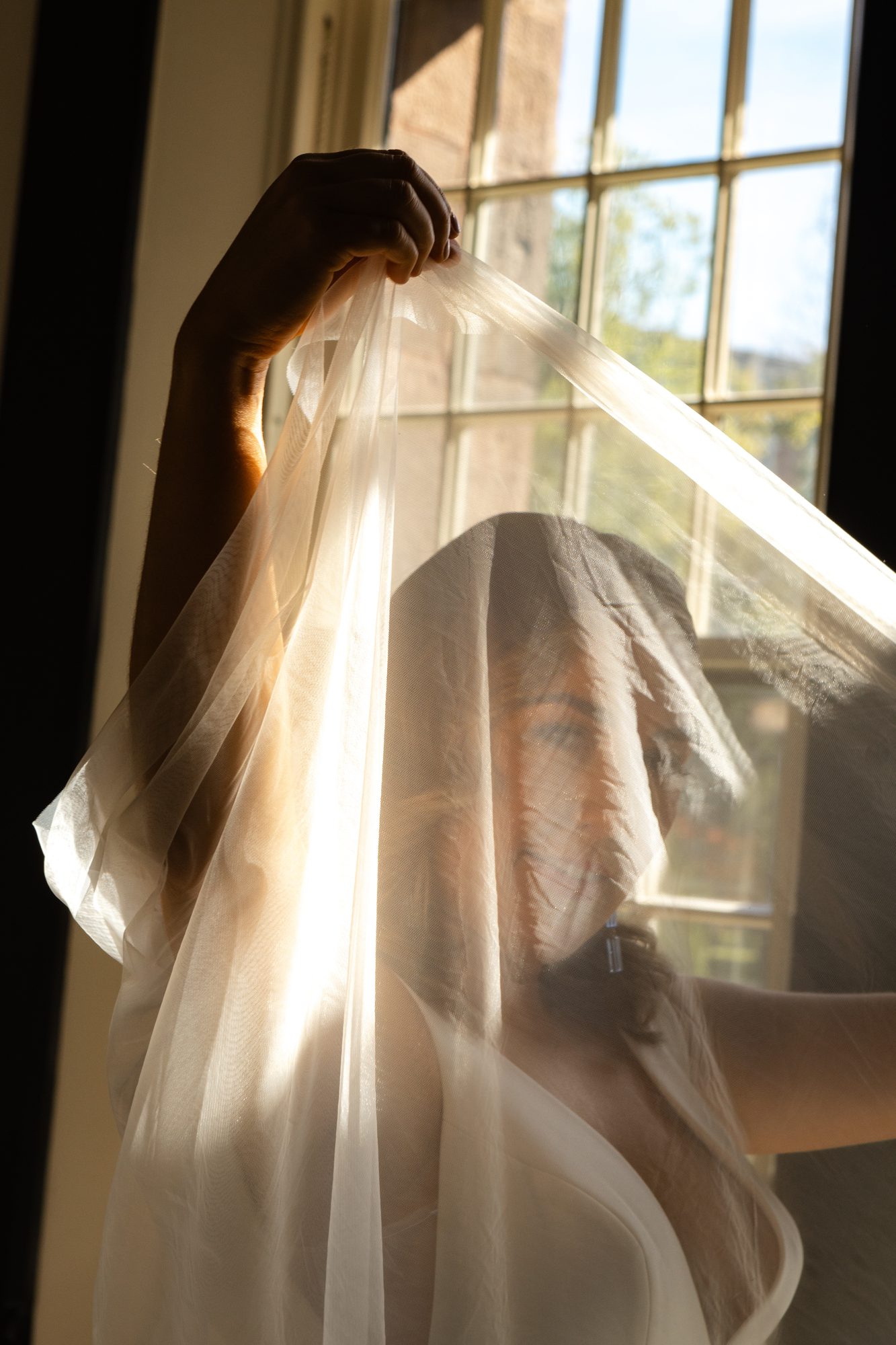 A hand holding a veil against a sunlit window, the bride's silhouette visible through the sheer fabric.