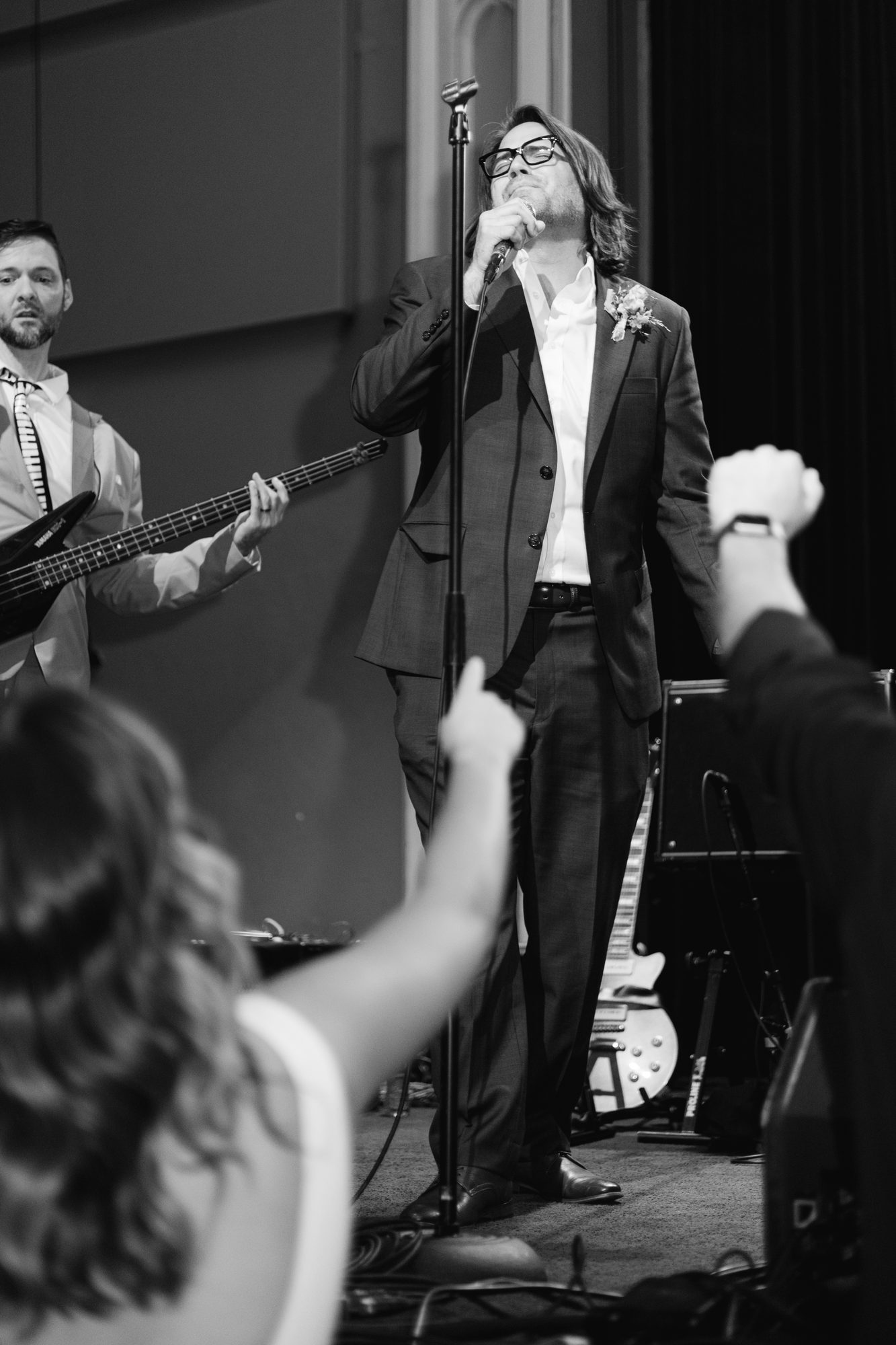 A groom singing into a microphone with the wedding band at the reception, guests reaching up.
