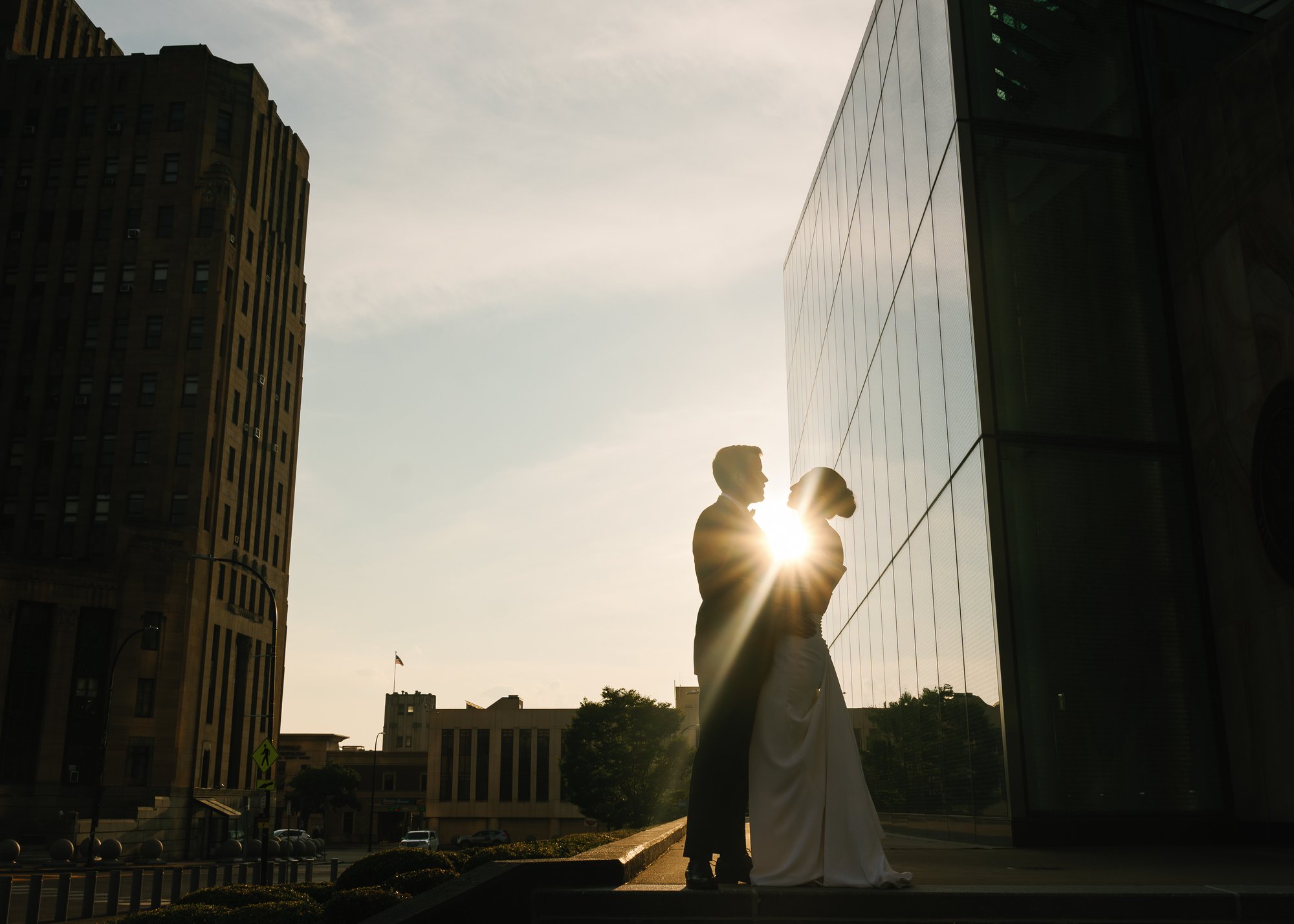A bride and groom silhouetted between city buildings at sunset, the sun breaking between them, downtown skyline visible behind.