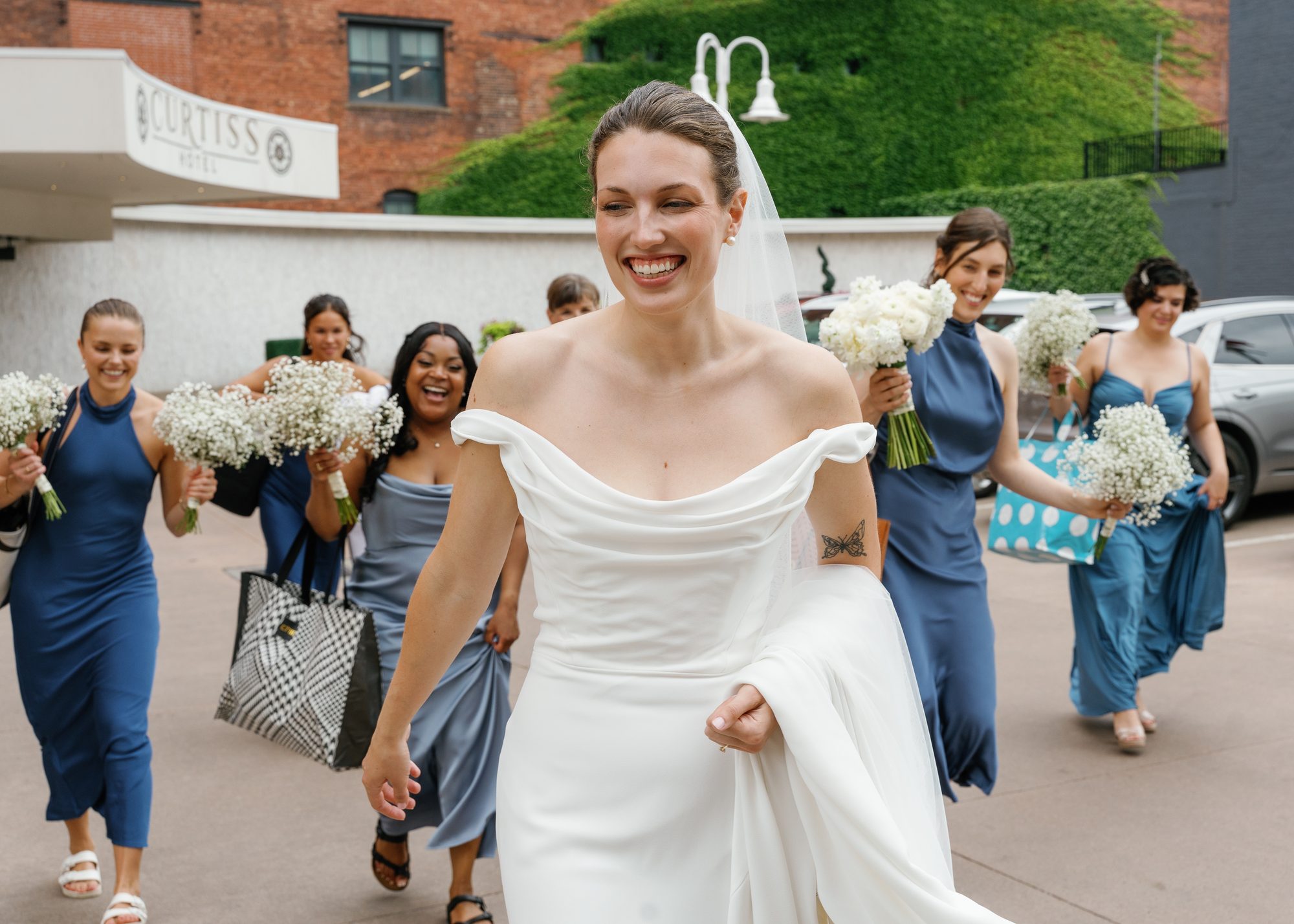 A bride laughing while walking with her bridesmaids in blue dresses outside a downtown hotel.