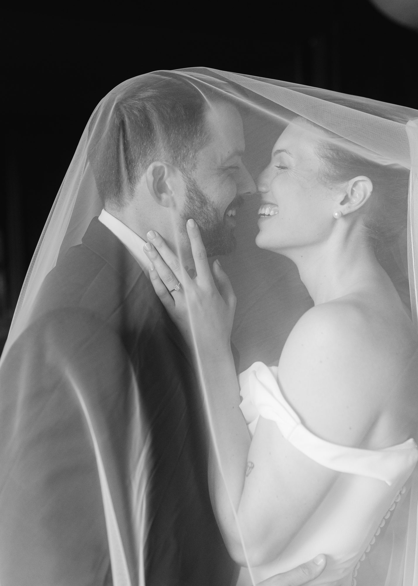 A bride and groom laughing together under a veil, foreheads close, black and white.