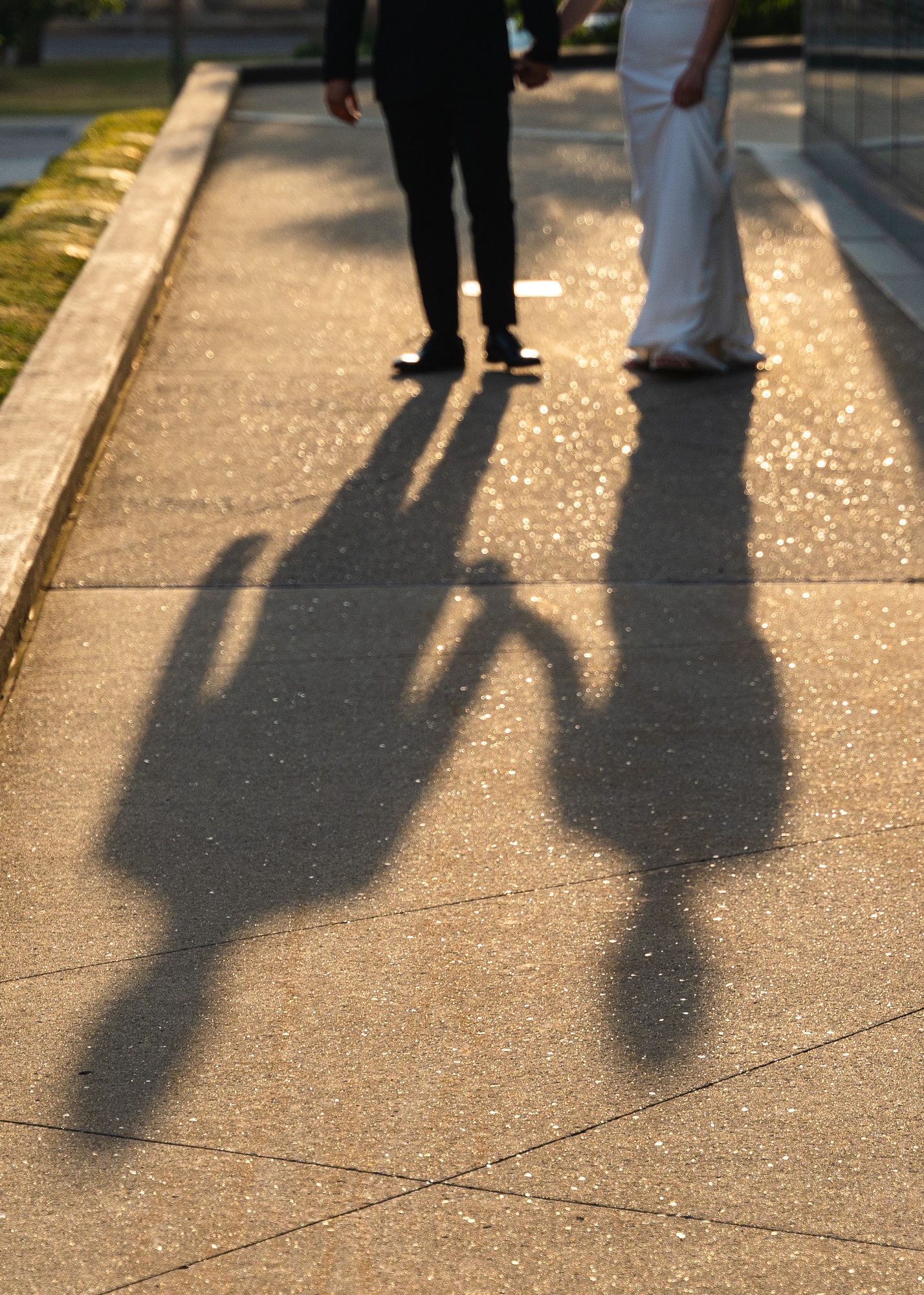 The long evening shadows of a bride and groom holding hands, cast across a sparkling sidewalk.