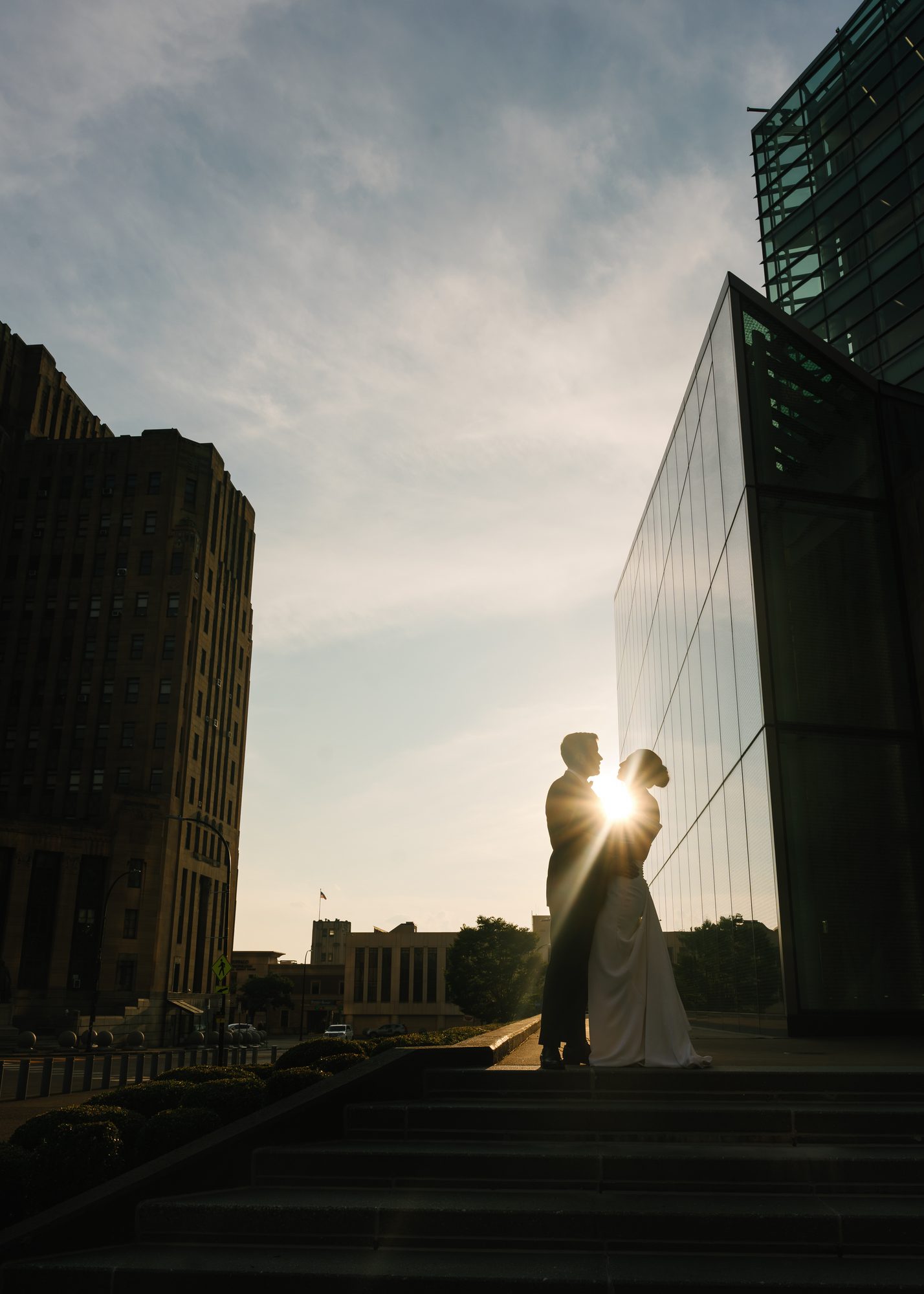 A bride and groom silhouetted between two glass buildings at sunset.