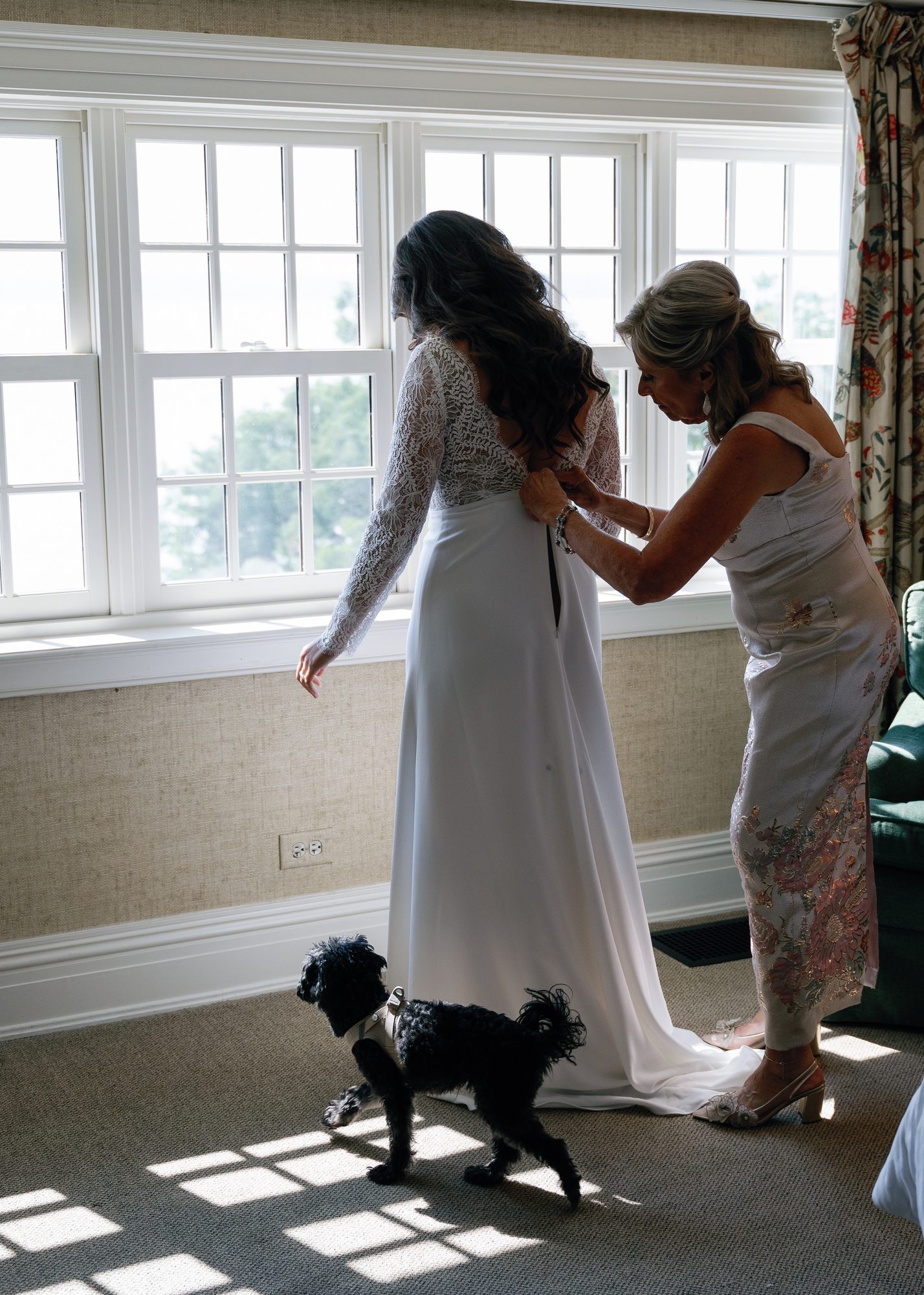 A bride having her dress laced up by her mother while a small dog walks across the sunlit floor.