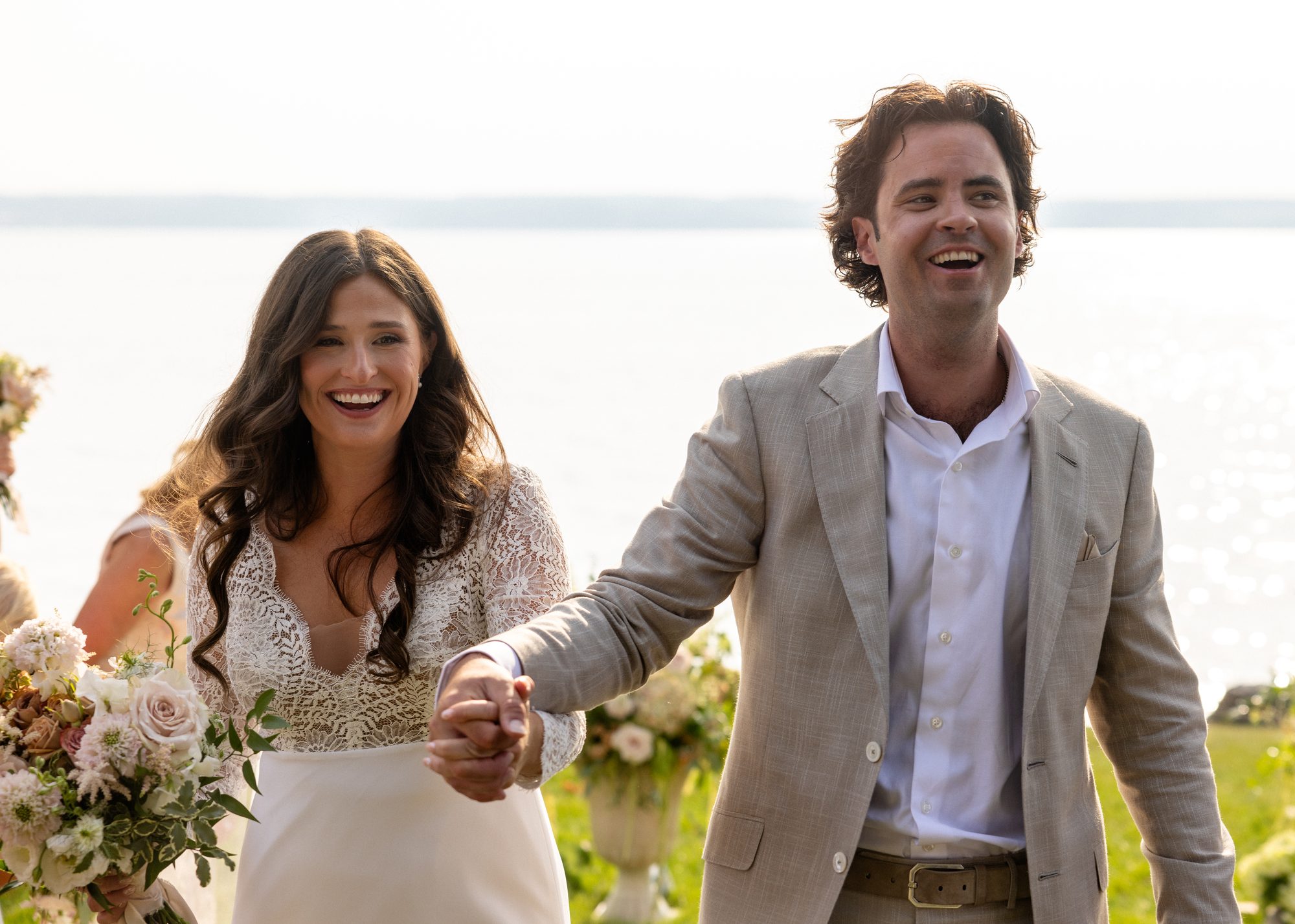 A bride and groom laughing while walking beside a lake in golden light.