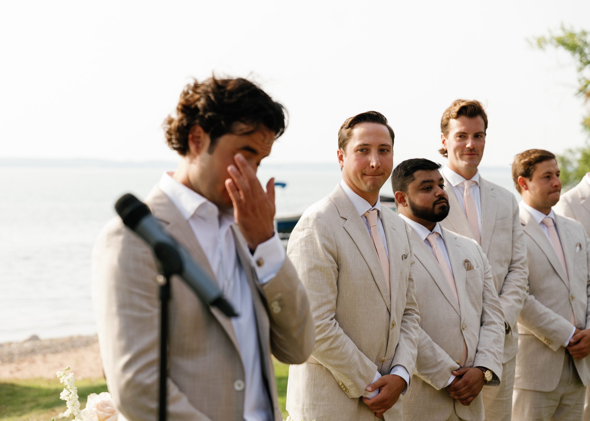 A groomsman wiping his eye during the ceremony while others in tan suits look on, lake in the background.