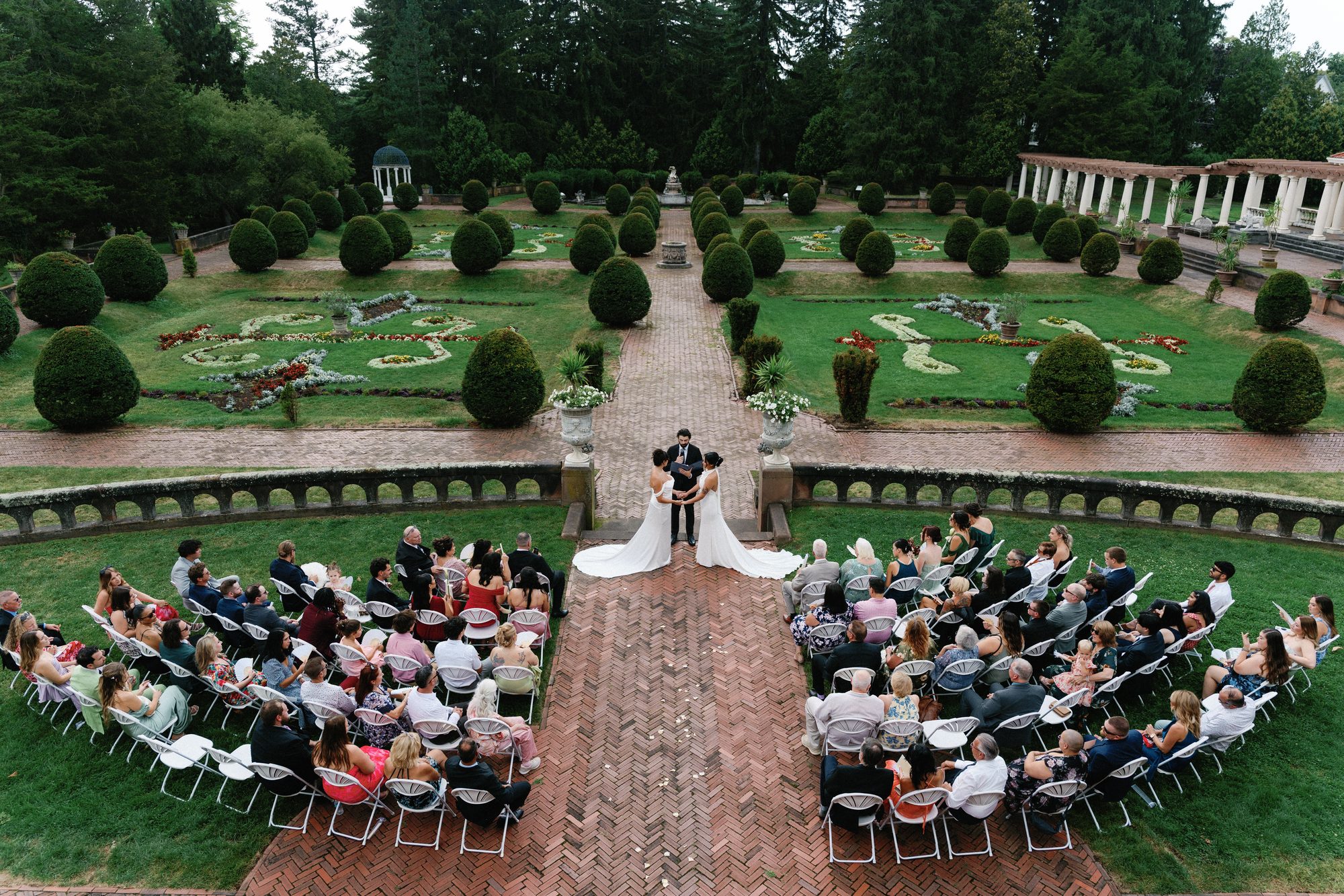 An aerial view of a formal garden estate wedding, two brides at the altar surrounded by topiary gardens and guests seated in a symmetric semi-circle.