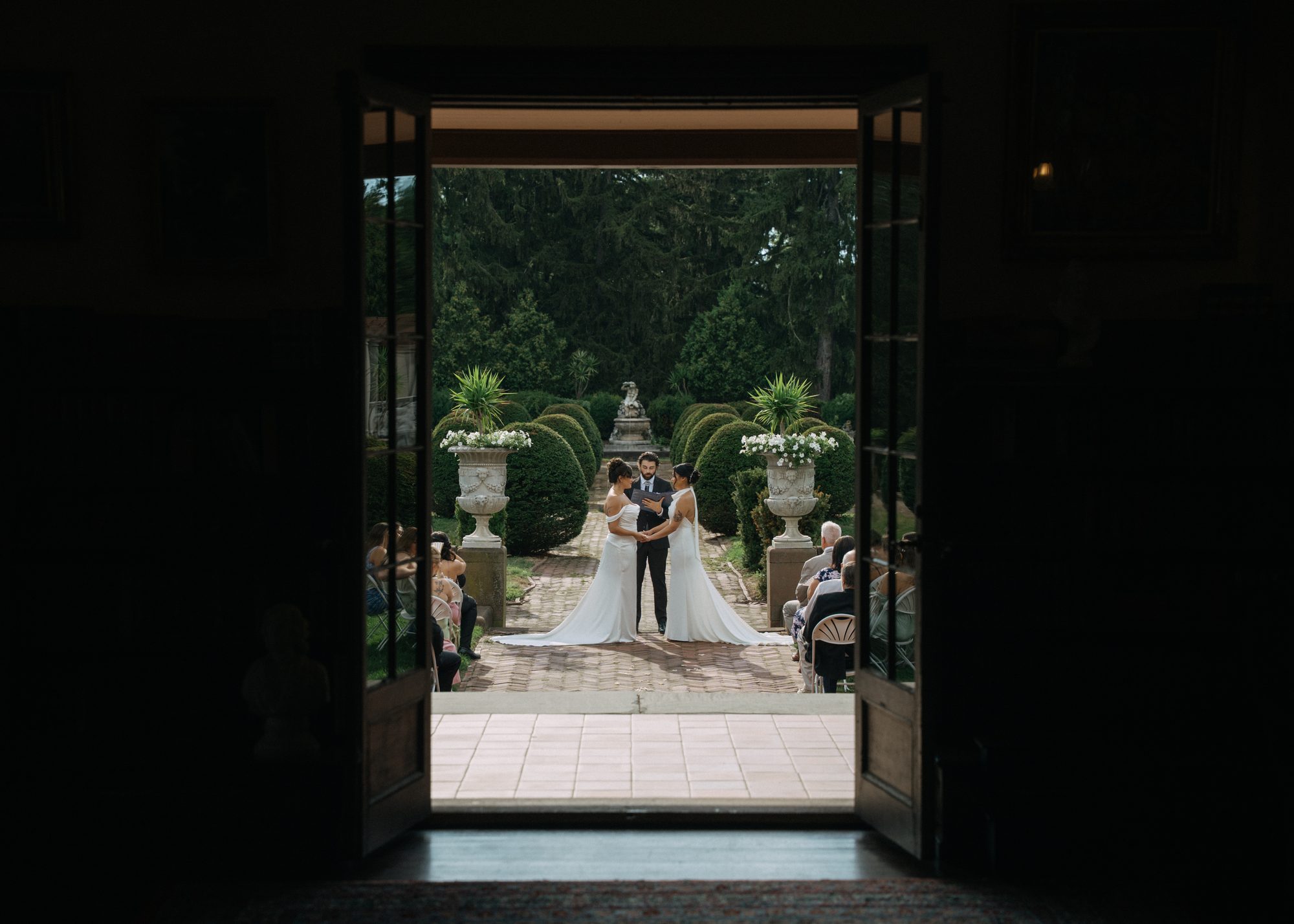 A garden ceremony framed through open double doors from inside a house, a couple and their officiant visible down the garden path.
