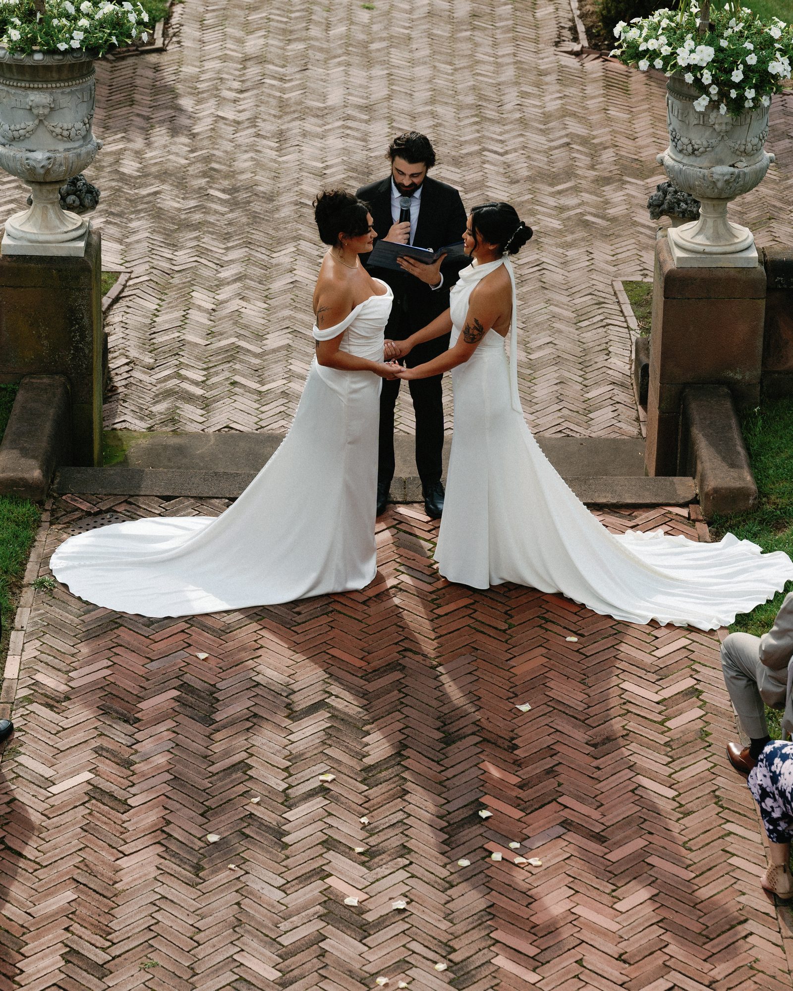 Two brides holding hands during their vows, an officiant reading between them, white petunias in stone urns on either side.
