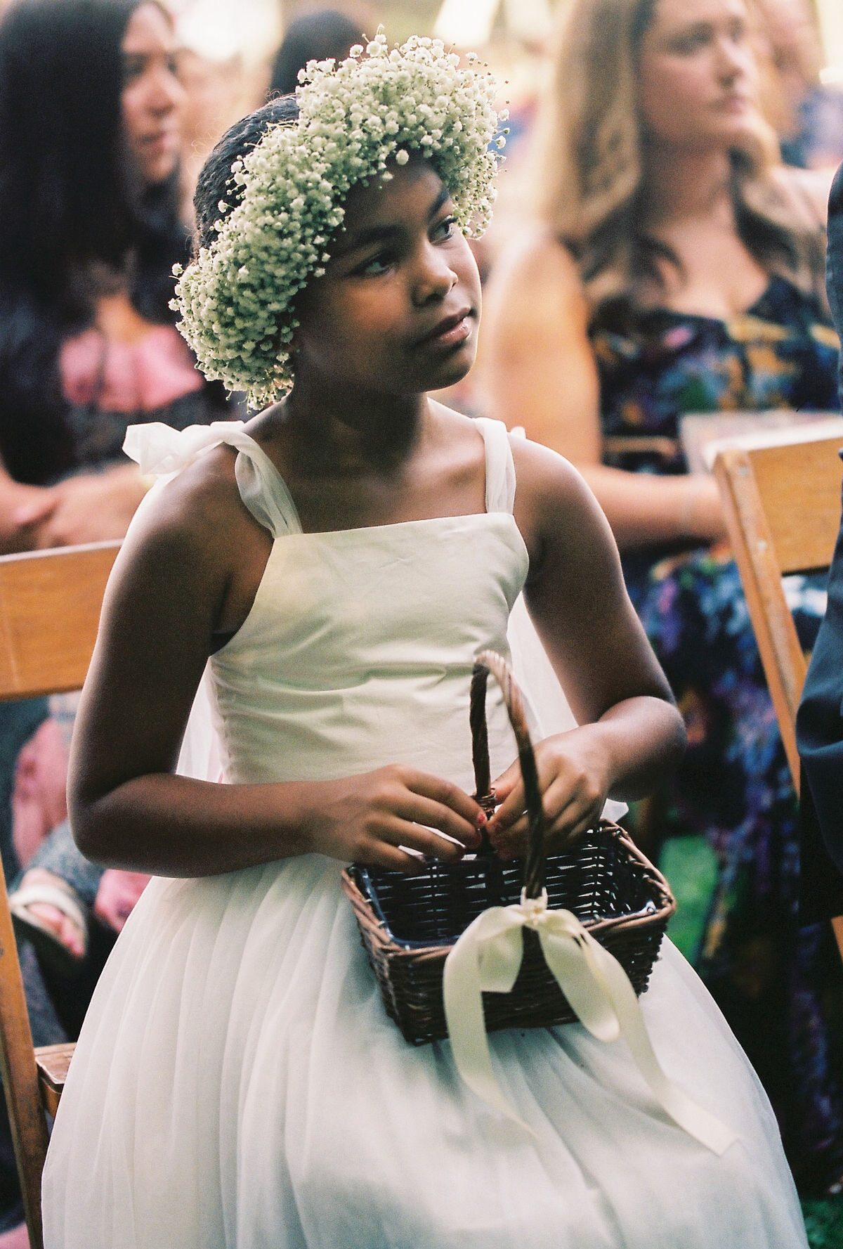 A young flower girl wearing a crown of baby's breath, holding a basket, looking off to the side during the ceremony.