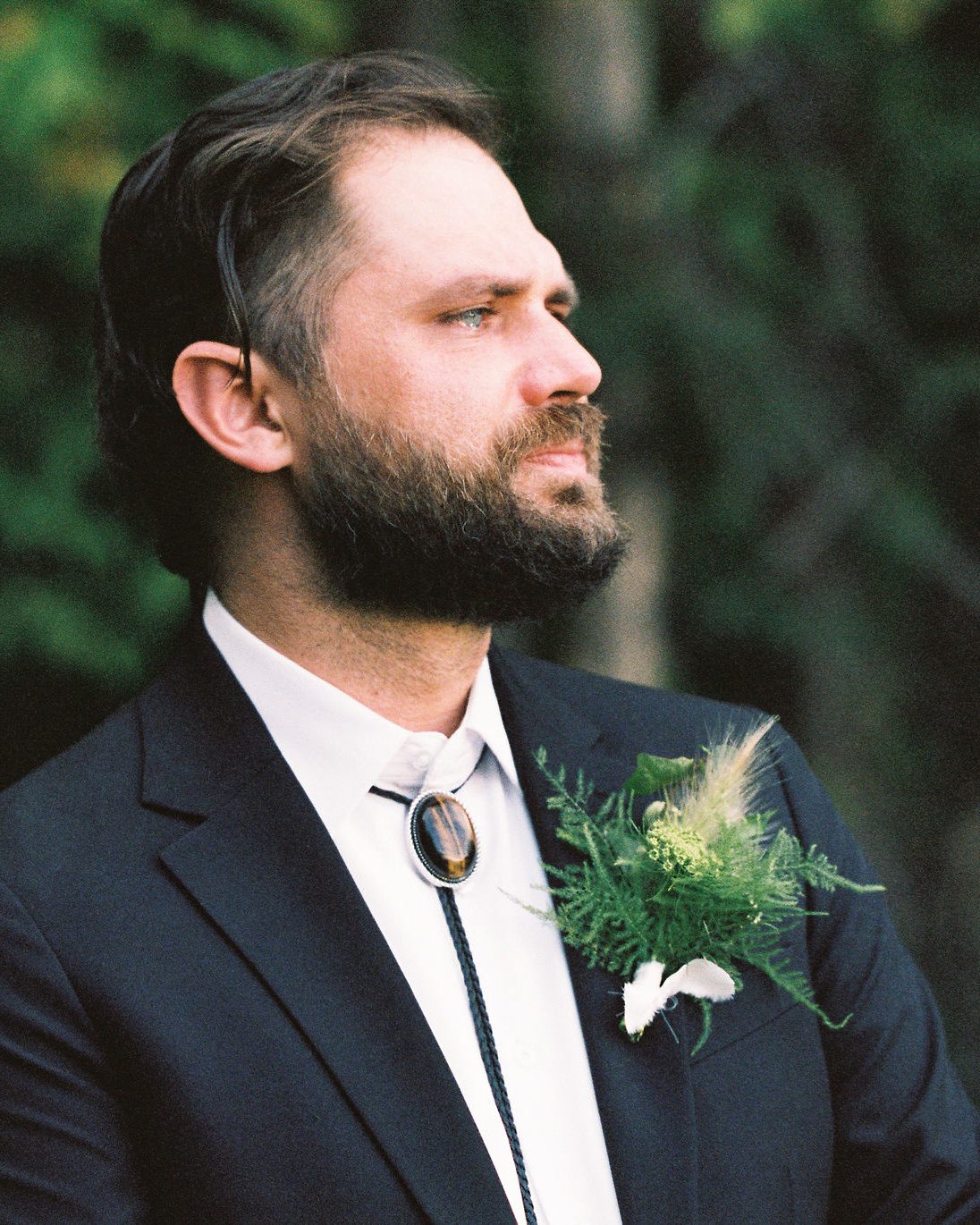 A groom in profile wearing a bolo tie and a fern boutonniere, looking off camera with a serious expression.