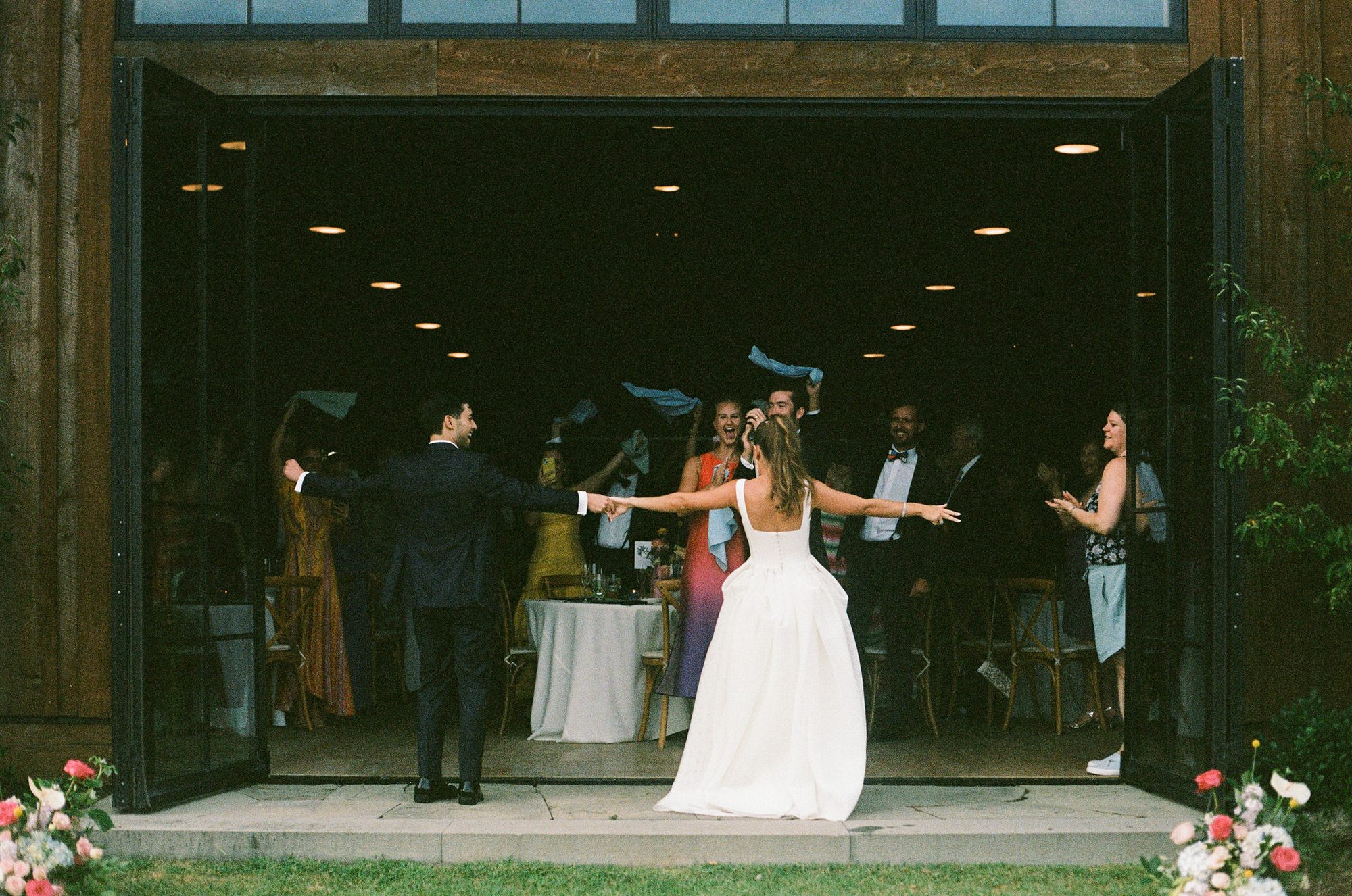 A couple entering their barn reception through open doors as guests cheer and wave napkins.