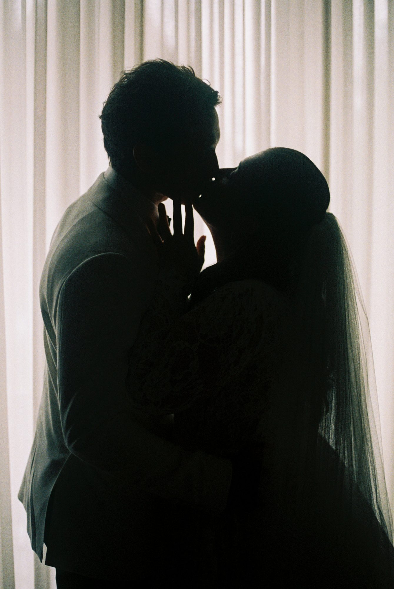 A bride and groom kissing, silhouetted against sheer curtains.