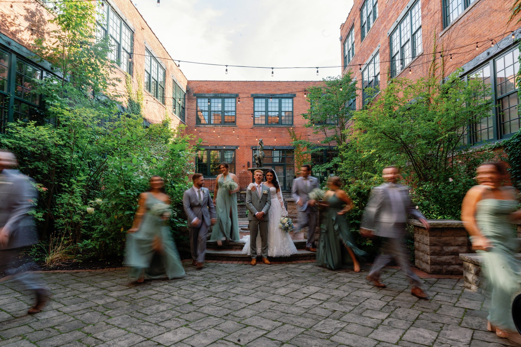 A bride and groom standing still at the center of a brick courtyard while their wedding party blurs past around them in motion.