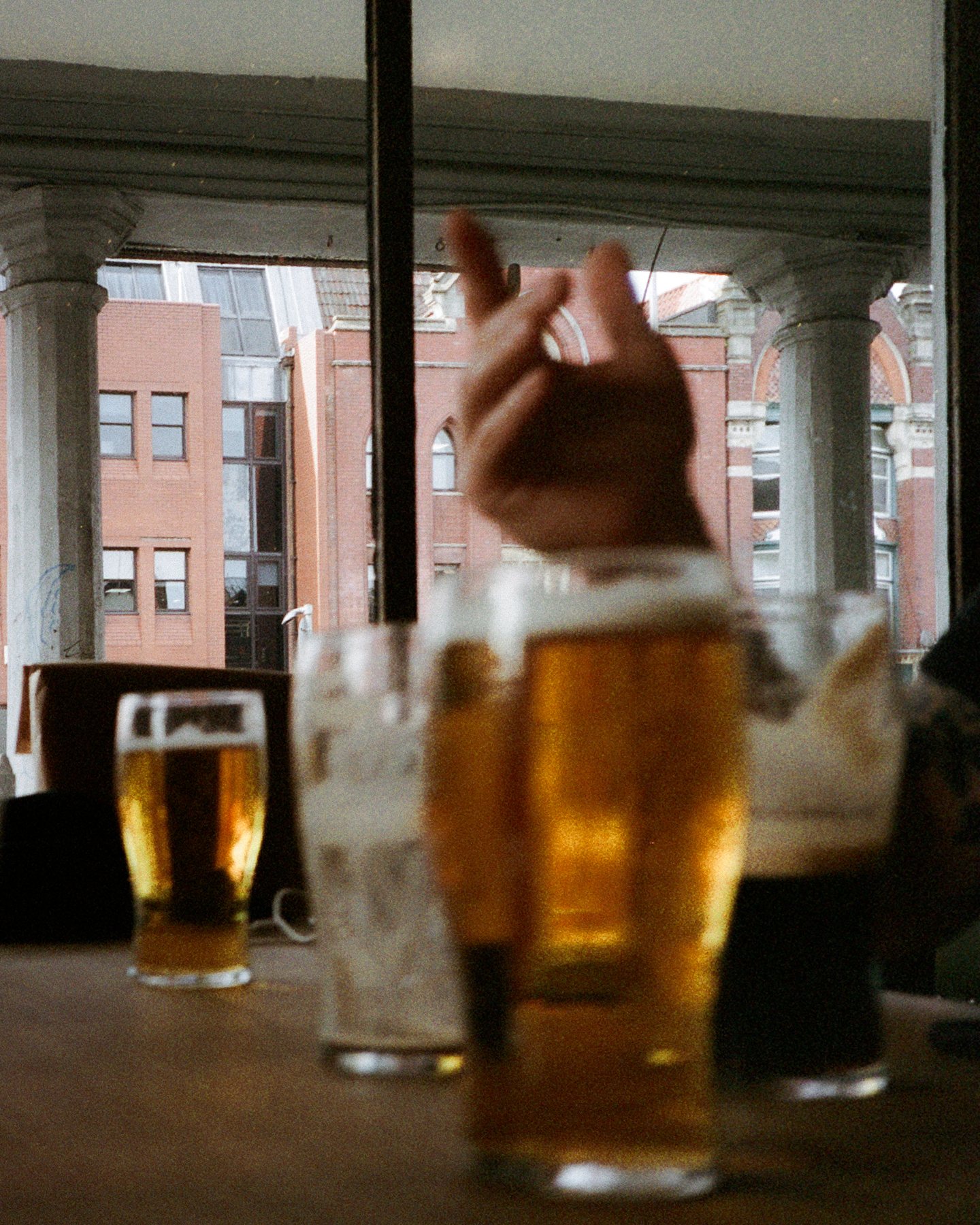 Glasses of beer on a table in front of a window, a hand reaching into frame, brick buildings visible outside.