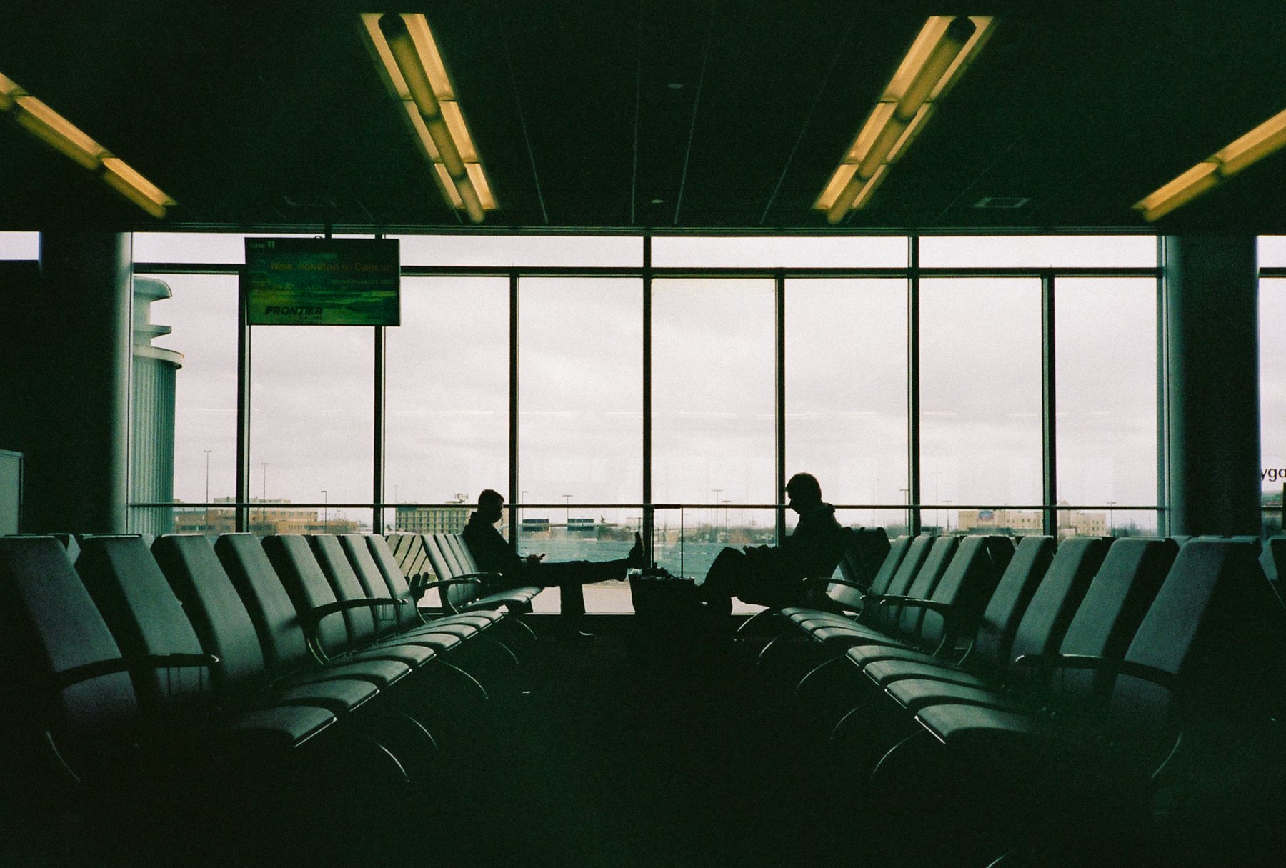 Two figures silhouetted in airport lounge seating, backlit by a wide bank of windows overlooking the tarmac at dusk.